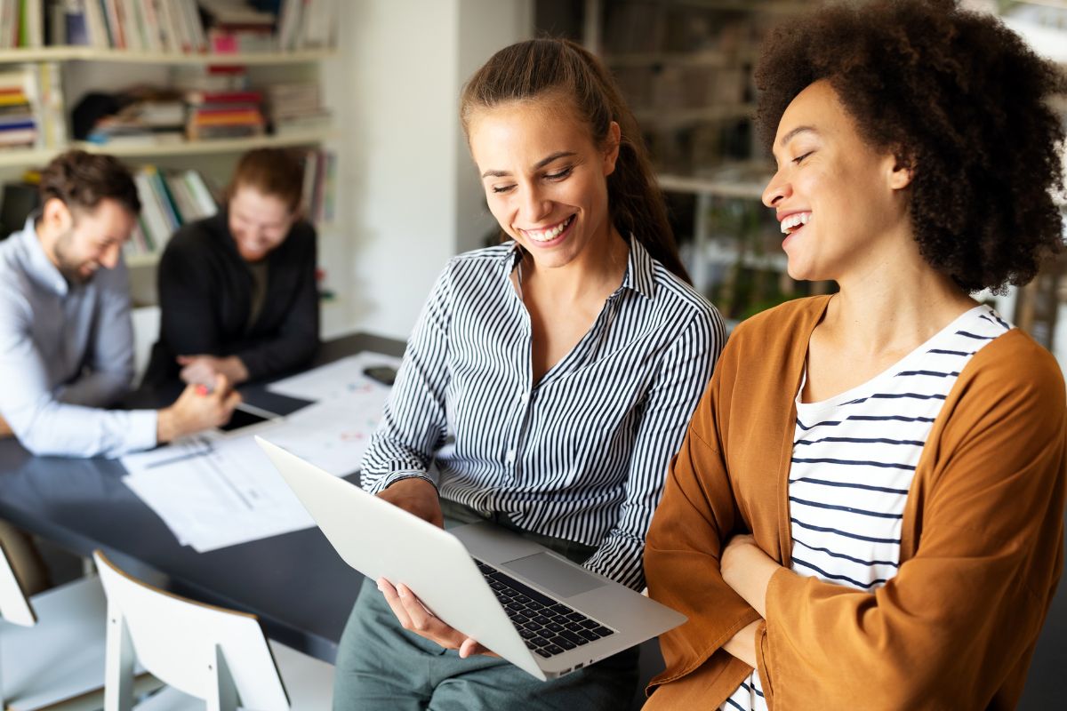 Two women smiling and looking at a laptop in the foreground, showing How To Use ChatGPT, with two men engaged in discussion in the background, sitting at a table with papers and notebooks in an office setting.