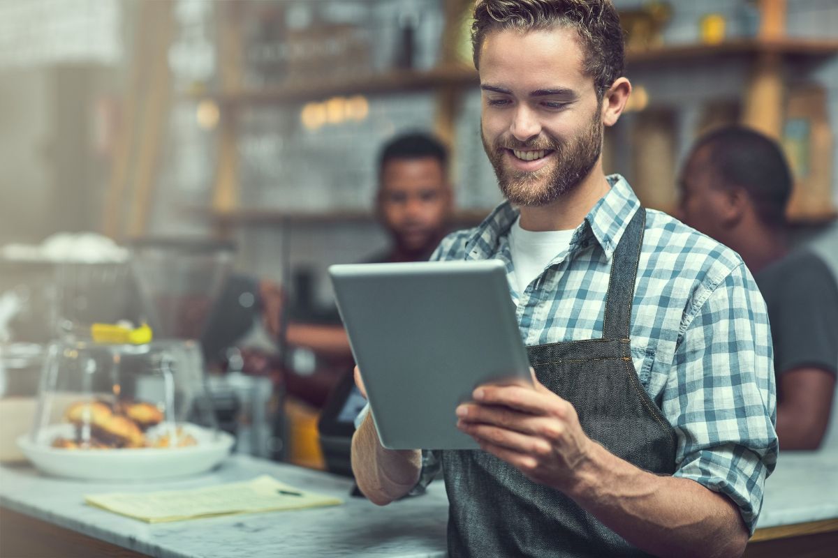 A barista wearing an apron holds a tablet and smiles in a coffee shop, showcasing how to use social media for business. Other staff and baked goods are visible in the background.