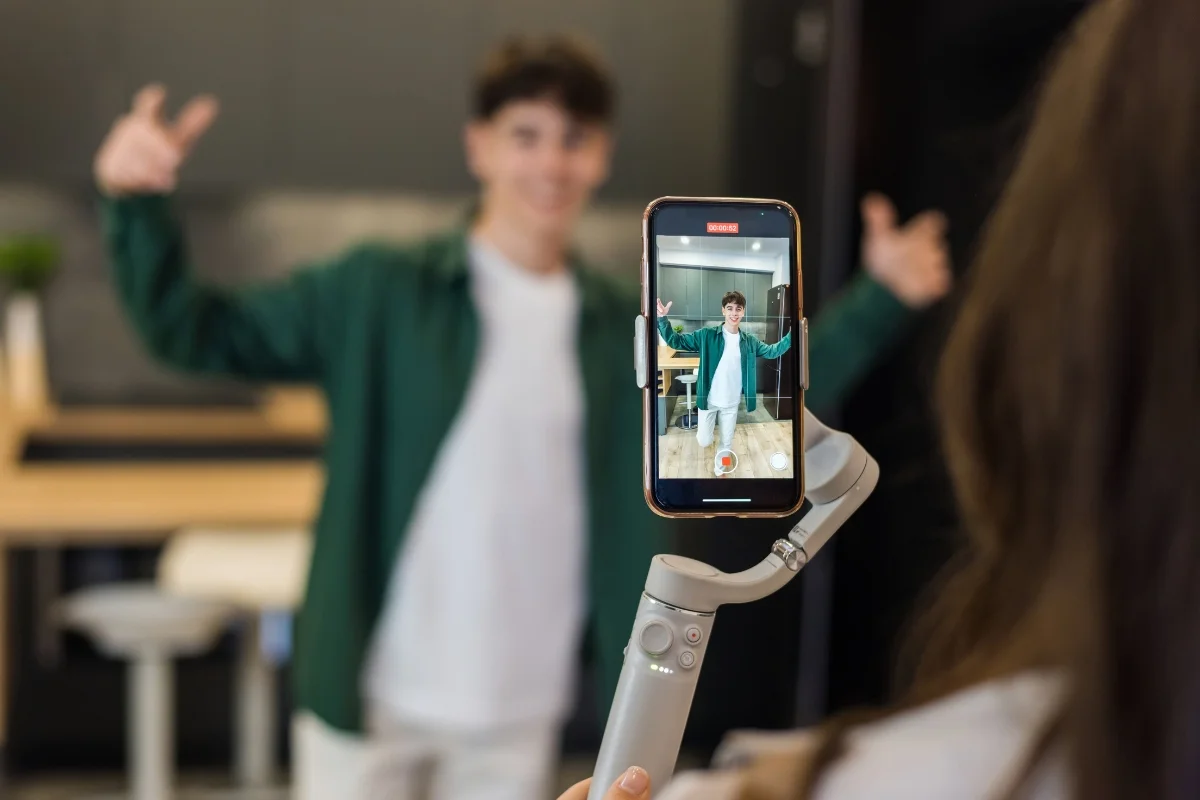 A person films another using a smartphone mounted on a handheld gimbal. The person being filmed, a local influencer, poses with both arms raised, capturing content for their marketing campaign.