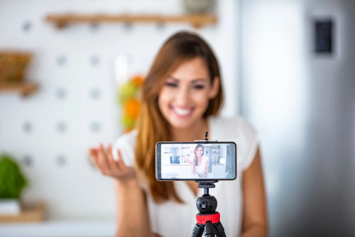 A local influencer is recording a video with a smartphone mounted on a tripod, smiling and gesturing with one hand in a brightly lit room.