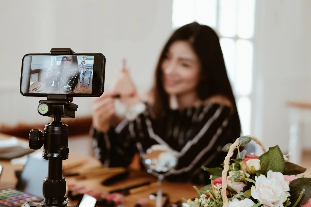 A woman smiles and holds a makeup brush during a live video recording session. A smartphone on a tripod captures the scene, perfect for local influencer marketing. Various beauty products and flowers are visible on the table, creating an inviting and professional setup.