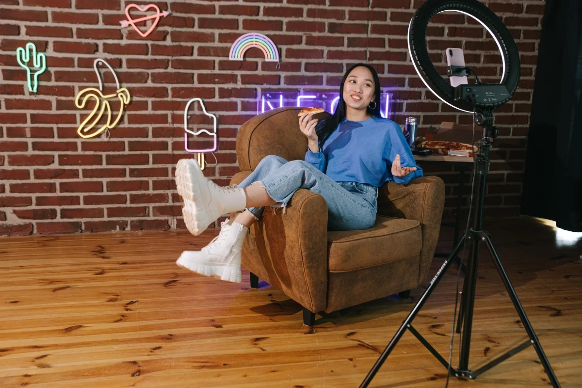 Person sitting on a brown armchair, holding a slice of pizza, while recording with a smartphone on a tripod in a room with neon signs and wooden flooring. The setup hints at their dive into local influencer marketing.