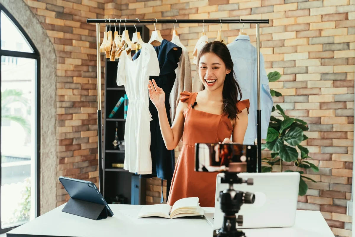 A woman stands in front of a clothing rack, waving at a smartphone on a tripod. Surrounded by an open laptop, a tablet, and a book, she's engaging in local influencer marketing to showcase the latest fashion trends.