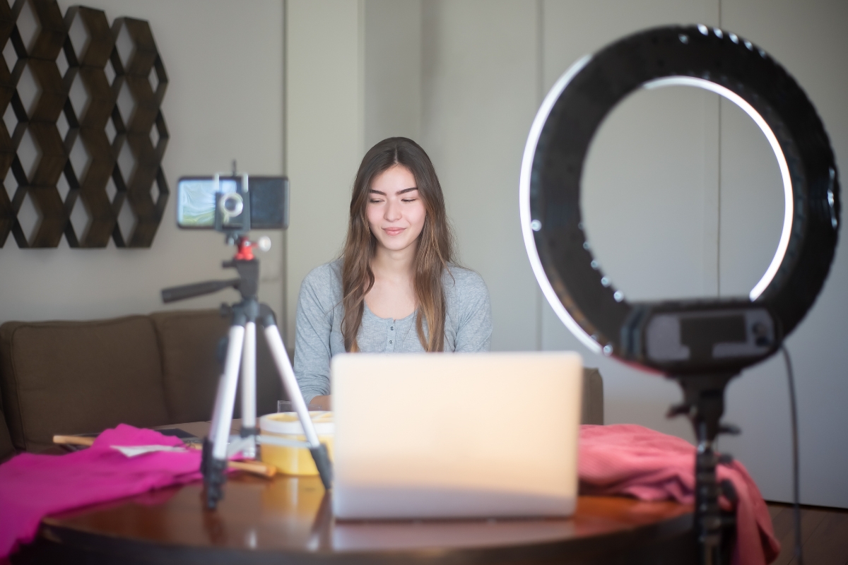 A woman sits at a table with a laptop, facing a smartphone on a tripod and a ring light, surrounded by various items, showcasing her dedication to local influencer marketing.