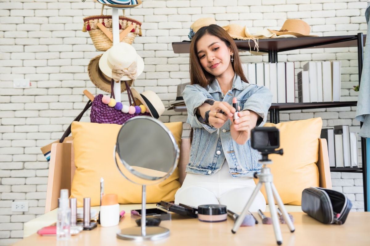 A woman in a denim jacket applies makeup at a table with various cosmetic products, while filming herself with a camera on a tripod. As part of her local influencer marketing strategy, shelves with hats and books are displayed in the background.