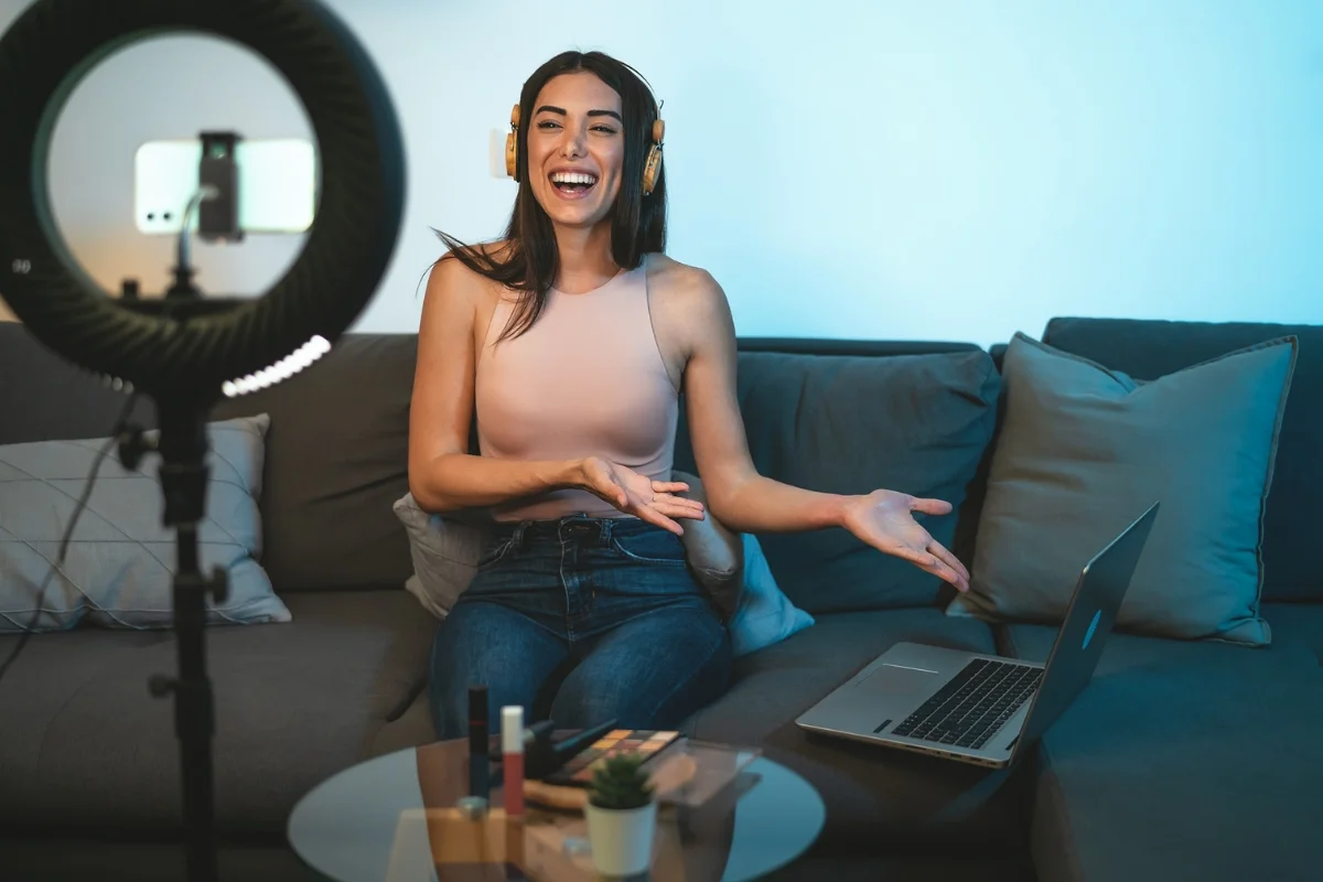 A woman wearing headphones sits on a couch, smiling and gesturing towards a laptop, with a ring light in front of her. Various items are on a round table in front of the couch, showcasing her skills in local influencer marketing.