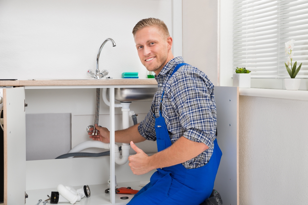 A plumber in blue overalls fixes pipes under a kitchen sink, giving a thumbs-up while kneeling. Tools and parts for plumbing automation are placed around him.