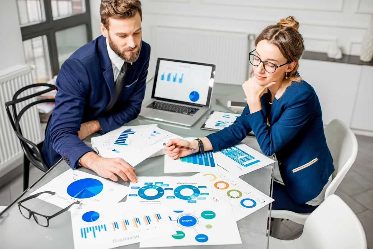 Two business professionals, specializing in plumbing automation, analyze charts and graphs at a desk with a laptop, wearing formal attire in a modern office setting.