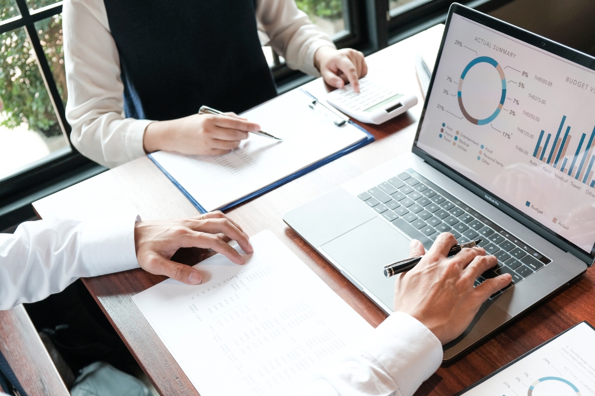 Two people working; one is using a calculator and writing on a clipboard, while the other is typing on a laptop displaying charts and graphs about plumbing automation. A document with relevant figures lies on the table near them.