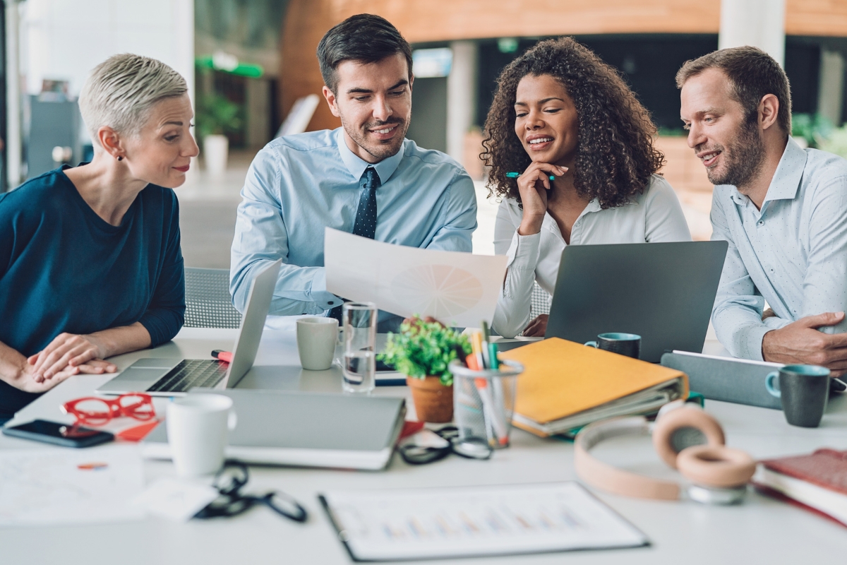 A diverse group of four colleagues sits at a desk, engaged in a meeting with laptops and documents. They are smiling and discussing something related to plumbing automation while looking at a piece of paper.