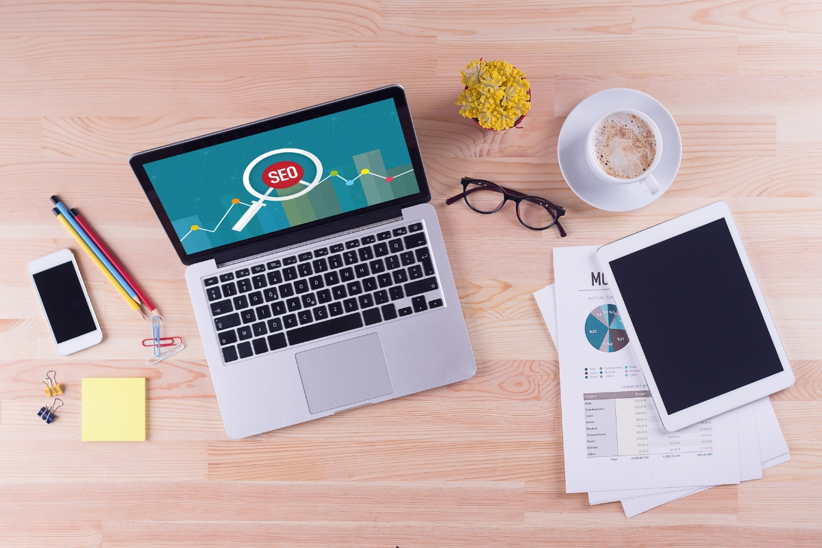 A laptop displaying an SEO graph, surrounded by a smartphone, colorful pencils, a yellow flower in a pot, reading glasses, service area pages documents, and a tablet on a wooden desk.