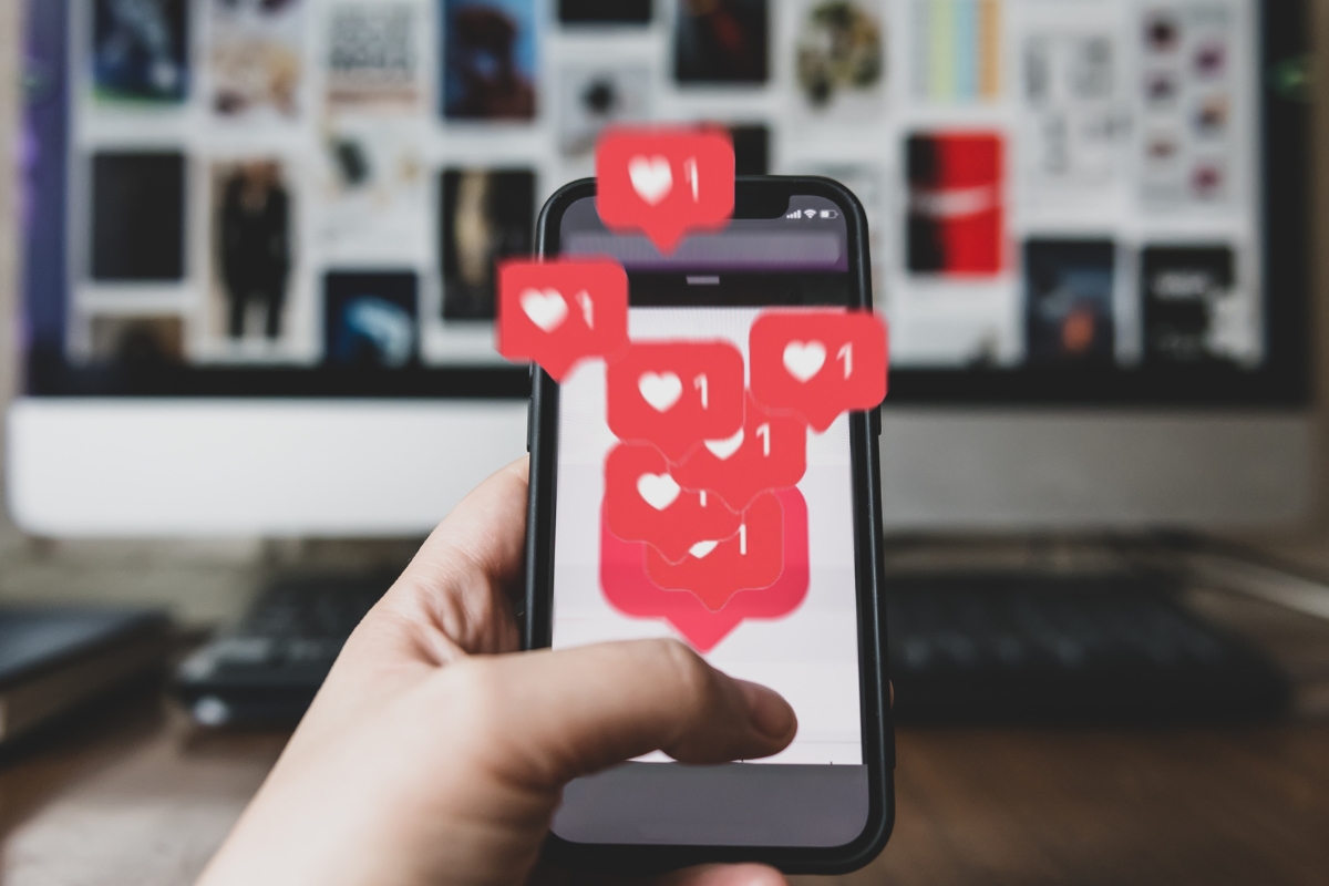 A hand holding a smartphone with multiple heart-shaped social media notifications appearing on the screen, showcasing social media marketing for local businesses. In the background, a computer monitor displays a grid of images.