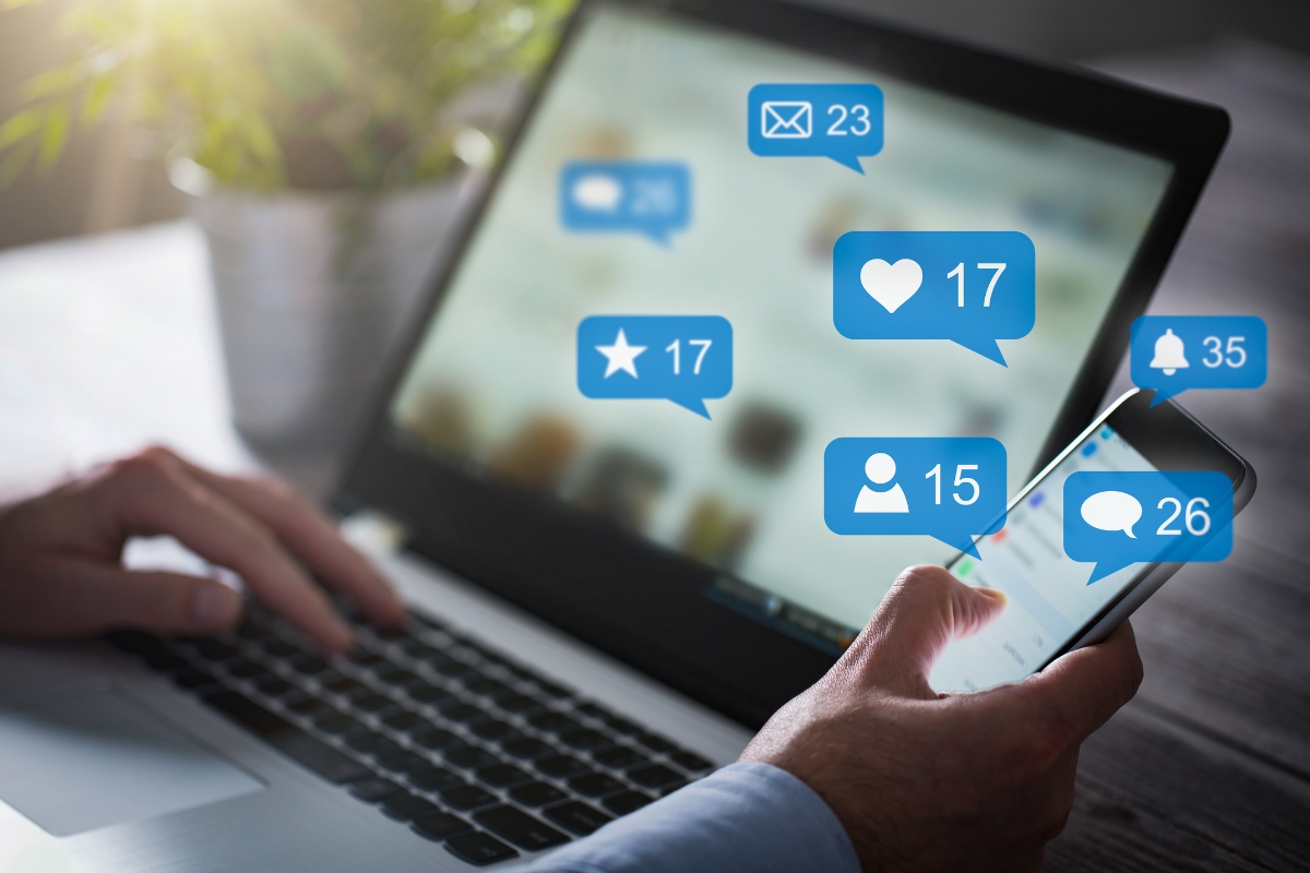 A person operates a laptop and holds a smartphone displaying social media notifications, including likes, messages, comments, and friend requests—perfect tools for effective social media marketing for local businesses.