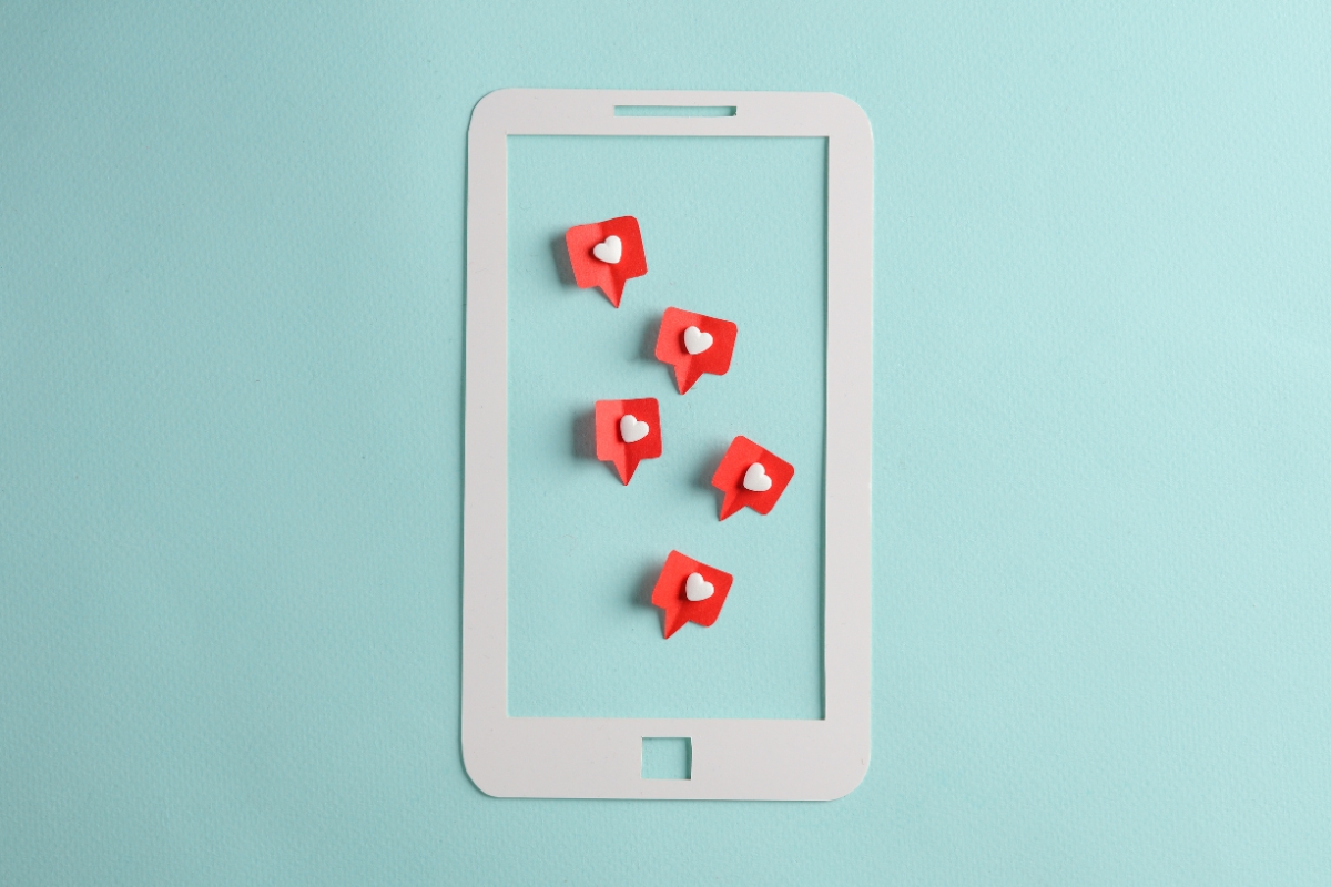 A white paper cutout of a smartphone with six red notification icons, each containing a white heart, sits on a light blue background, symbolizing the potential impact of social media marketing for local businesses.