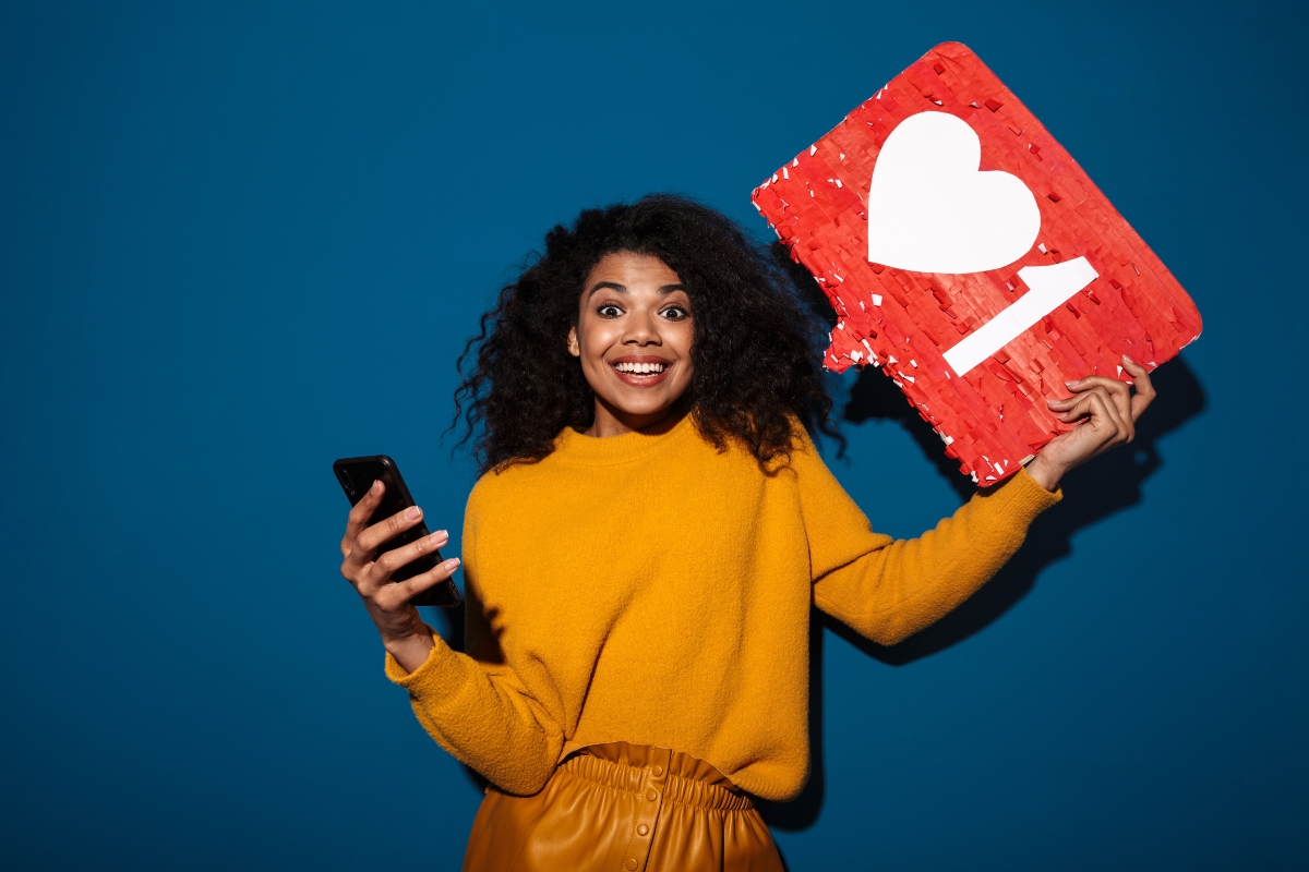 A woman in a yellow sweater holds a smartphone and a large red notification icon with a heart and the number one, against a blue background. She is smiling and appears happy, showcasing the power of social media marketing for local businesses.