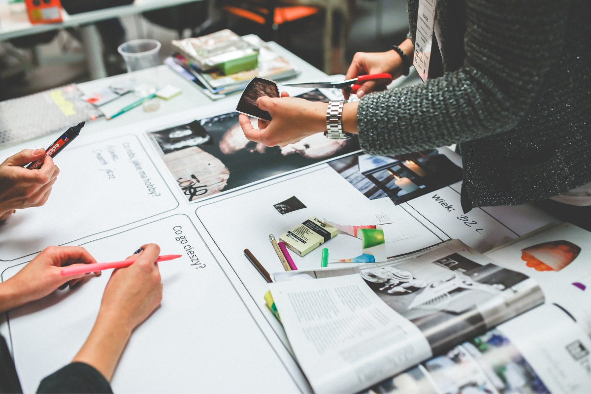 People working collaboratively at a table with notebooks, pens, and various supplies, including a smartphone, papers, craft materials, and a social media marketing plan.