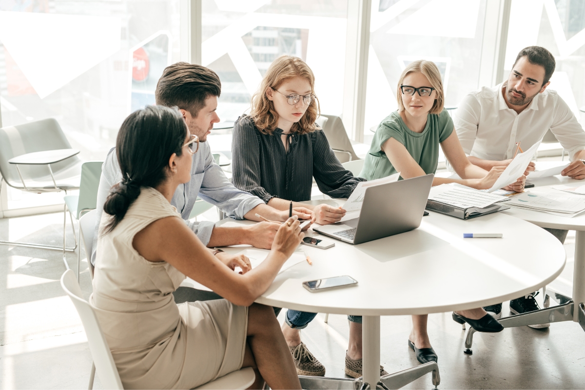 A group of five people in a meeting room are gathered around a table with a laptop, documents, and phones, engaged in a discussion about the social media marketing plan in a well-lit space with large windows.