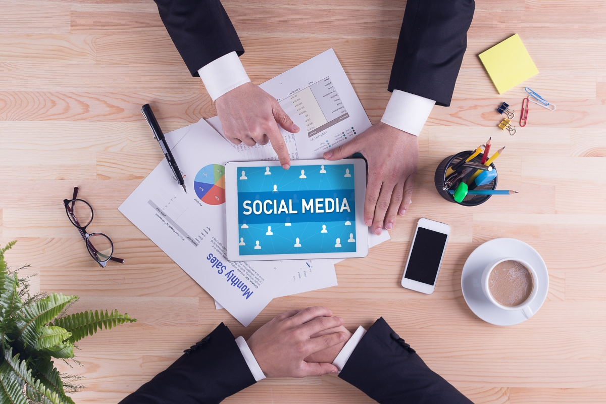 Three people in business attire around a wooden table are reviewing a social media marketing plan on a tablet. The table is adorned with papers, a phone, a potted plant, assorted stationery items, and a cup of coffee.