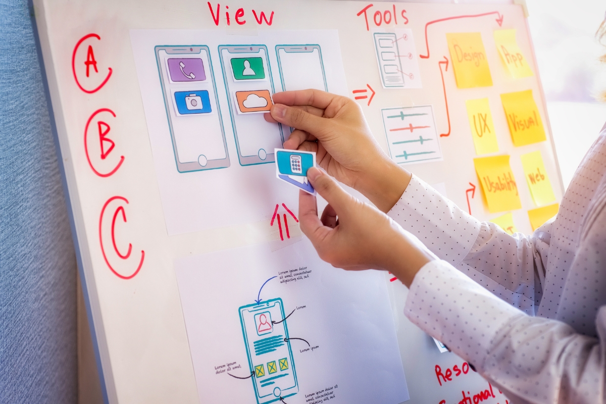 Person arranging app design mockups on a whiteboard, surrounded by notes and diagrams for a mobile app development project, focusing on user experience optimization.