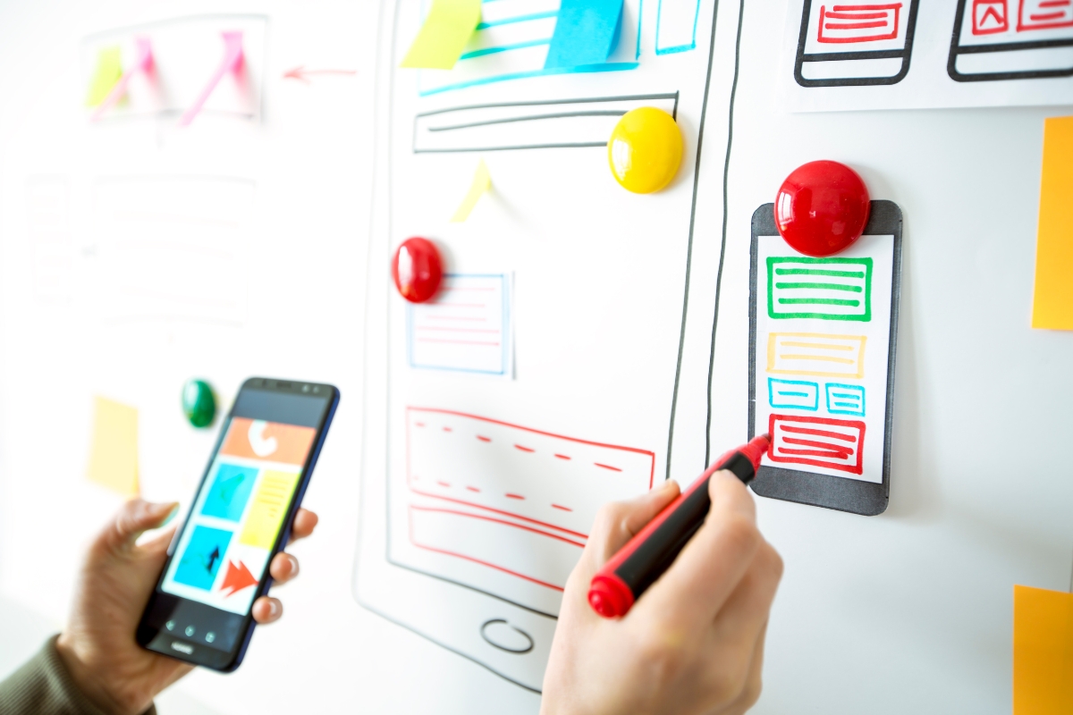 A person is holding a smartphone in their left hand and drawing a colorful website layout on a whiteboard with their right hand, surrounded by sticky notes and push pins, focusing on user experience optimization.