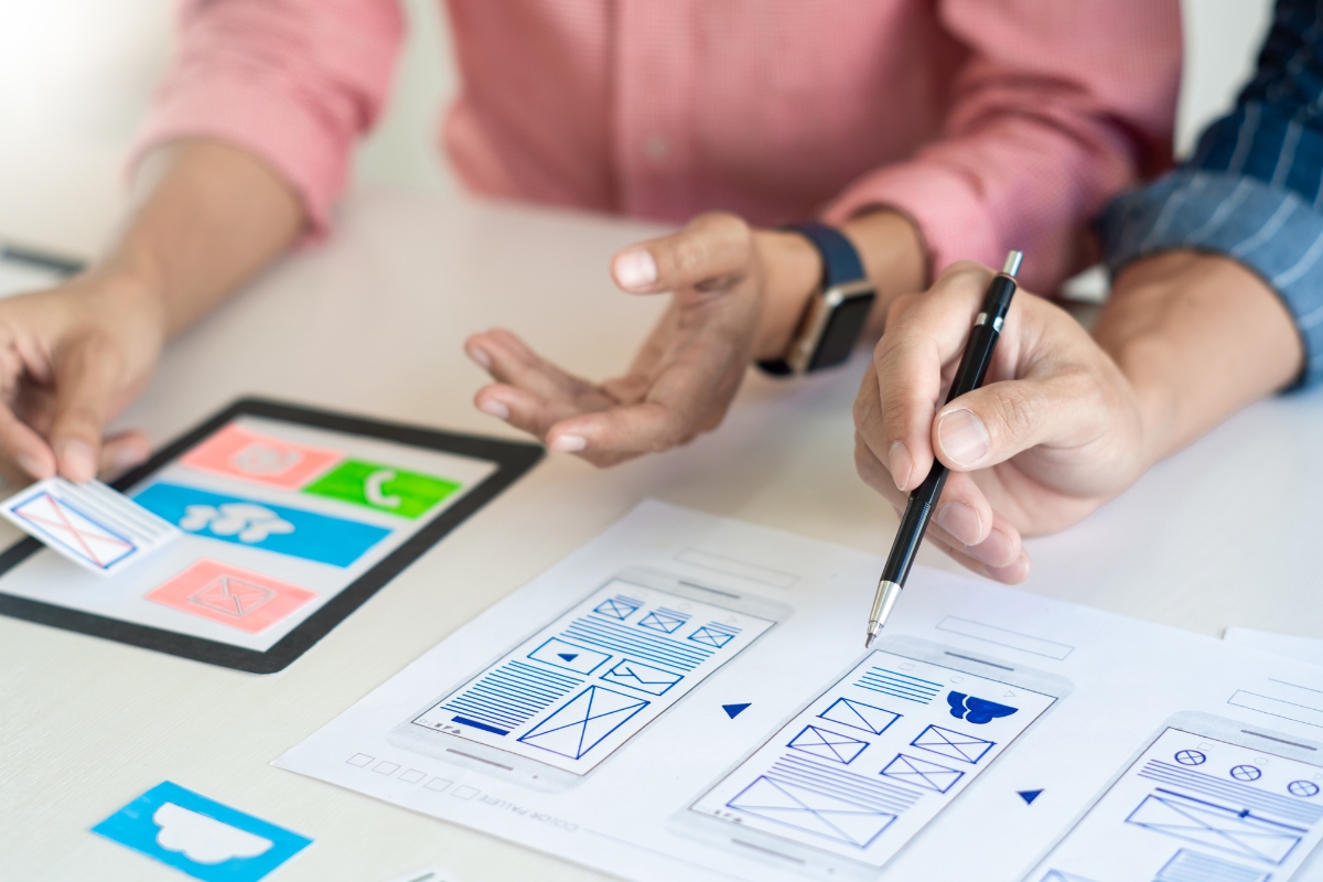 Two individuals are discussing and pointing at website wireframes and UI designs on a tablet and printed paper on a desk, focusing intently on user experience optimization.