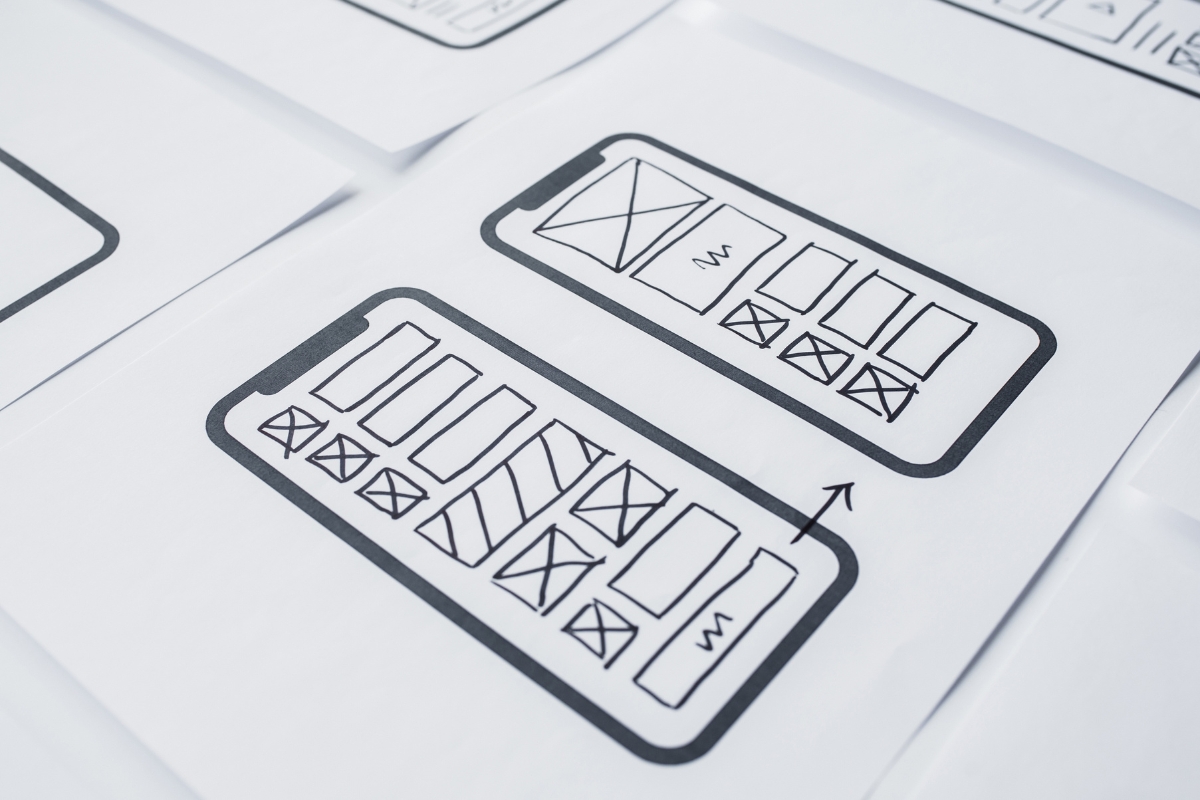 Papers displaying wireframe sketches of a mobile app interface layout, aimed at user experience optimization, are spread across a surface.