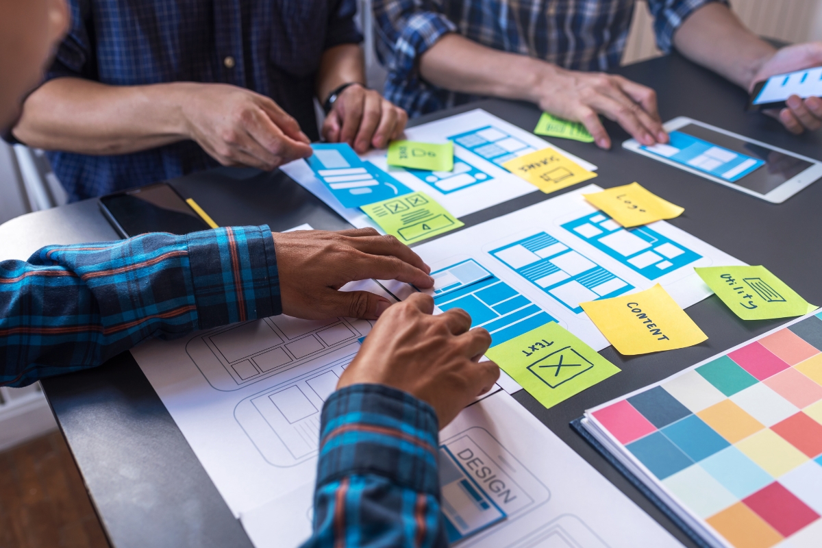 Three people collaborate on a design project, arranging paper prototypes and sticky notes labeled with content, design, and other elements on a table, all working toward user experience optimization.