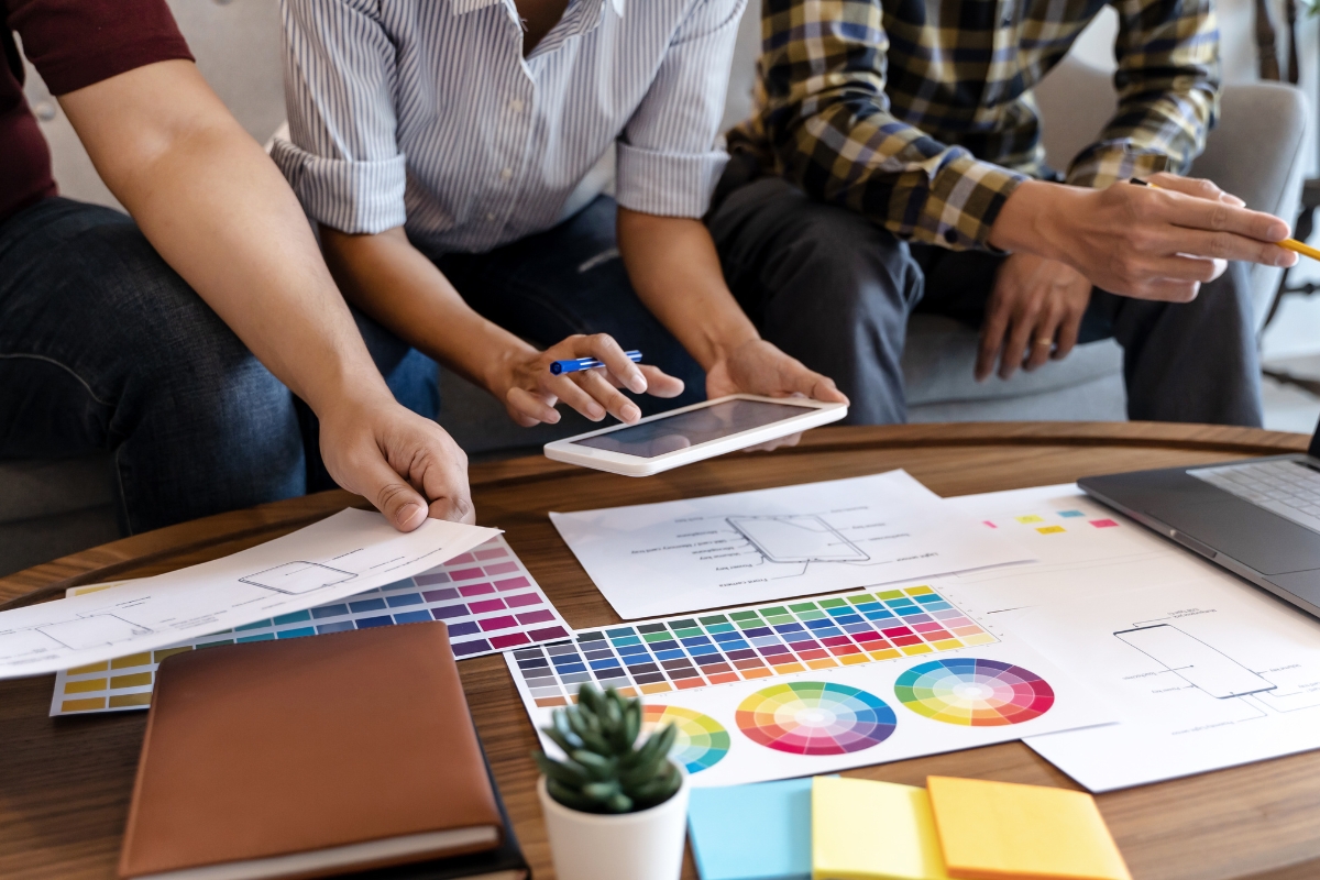 Three people working at a table with color swatches, design sketches, a tablet, and a laptop focused on user experience optimization. A small potted plant and a brown notebook are also on the table.