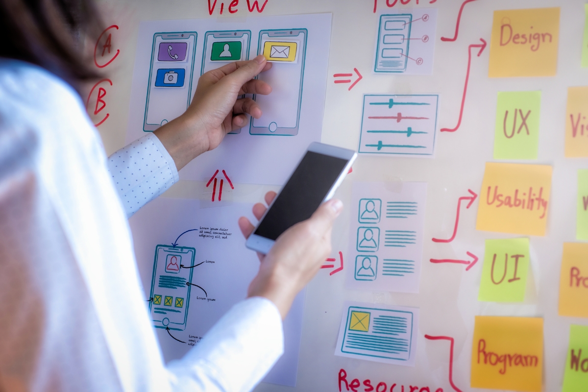 Person holding a smartphone and pointing to a design mockup on a whiteboard, which has various UI/UX elements and notes focused on user experience optimization.