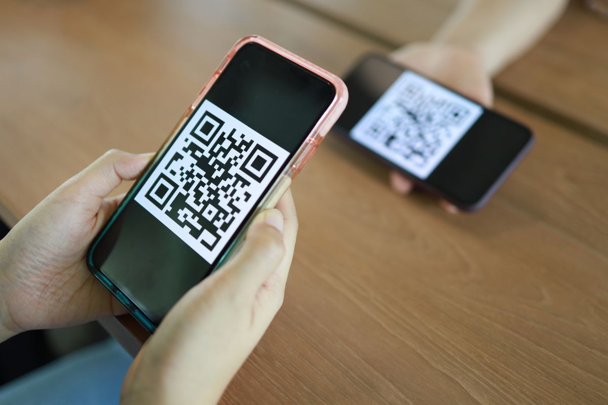 Two hands holding smartphones, each displaying a QR code on the screen. The phones are positioned over a wooden table, prompting viewers to wonder, "What are QR codes?