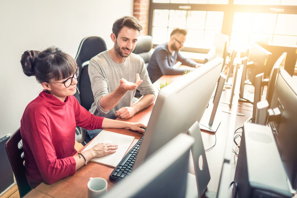 Two people are seated at desks, looking at computer screens and discussing something, possibly asking "What is NLP?" A third person in the background is also working on a computer. The room is well-lit with natural light.