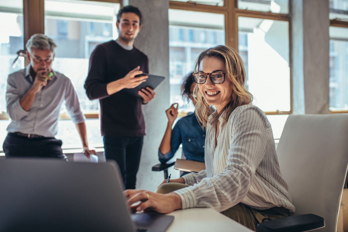 A woman wearing glasses smiles at her laptop in a bright office, likely reading an article on how to do keyword research. Three colleagues stand in the background, engaged in discussion.