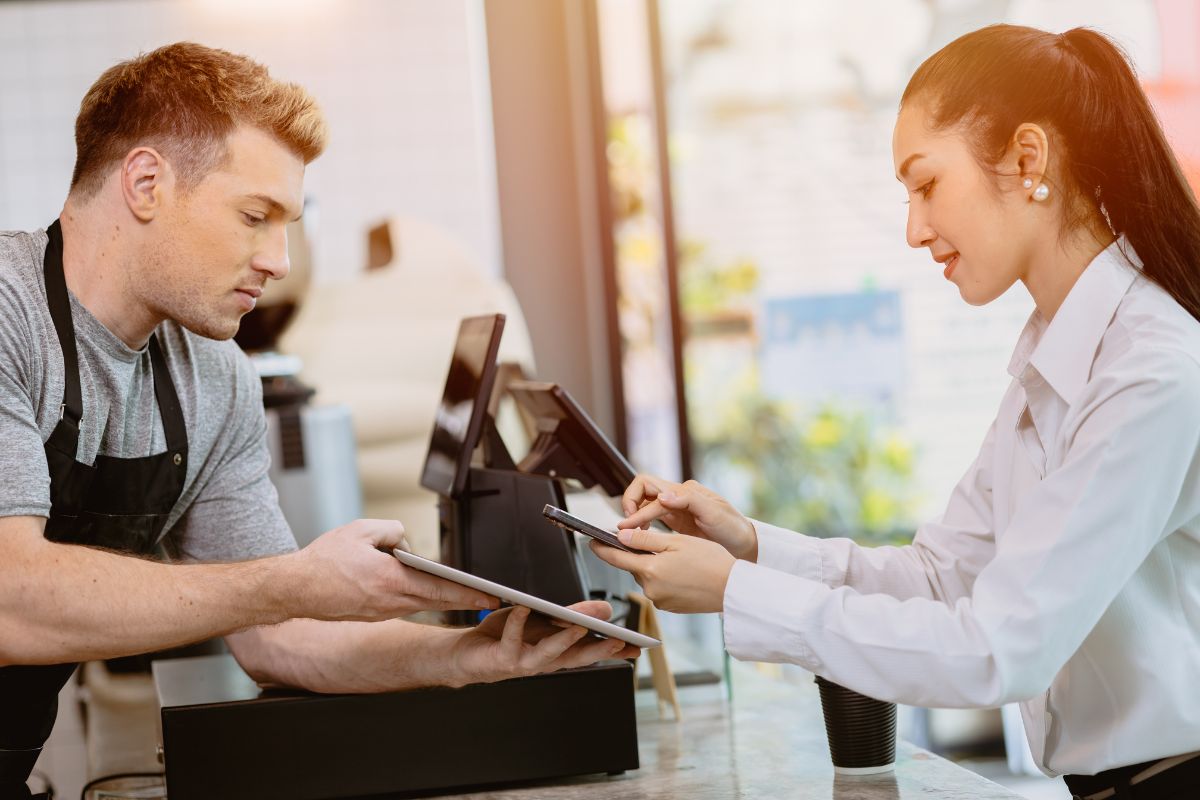 A customer uses a smartphone to make a payment to a barista behind the counter while holding a tablet. Both individuals, focused on the transaction, demonstrate modern convenience and efficiency. Curious observers might wonder, "What are QR codes?" as they notice one being scanned for the payment.