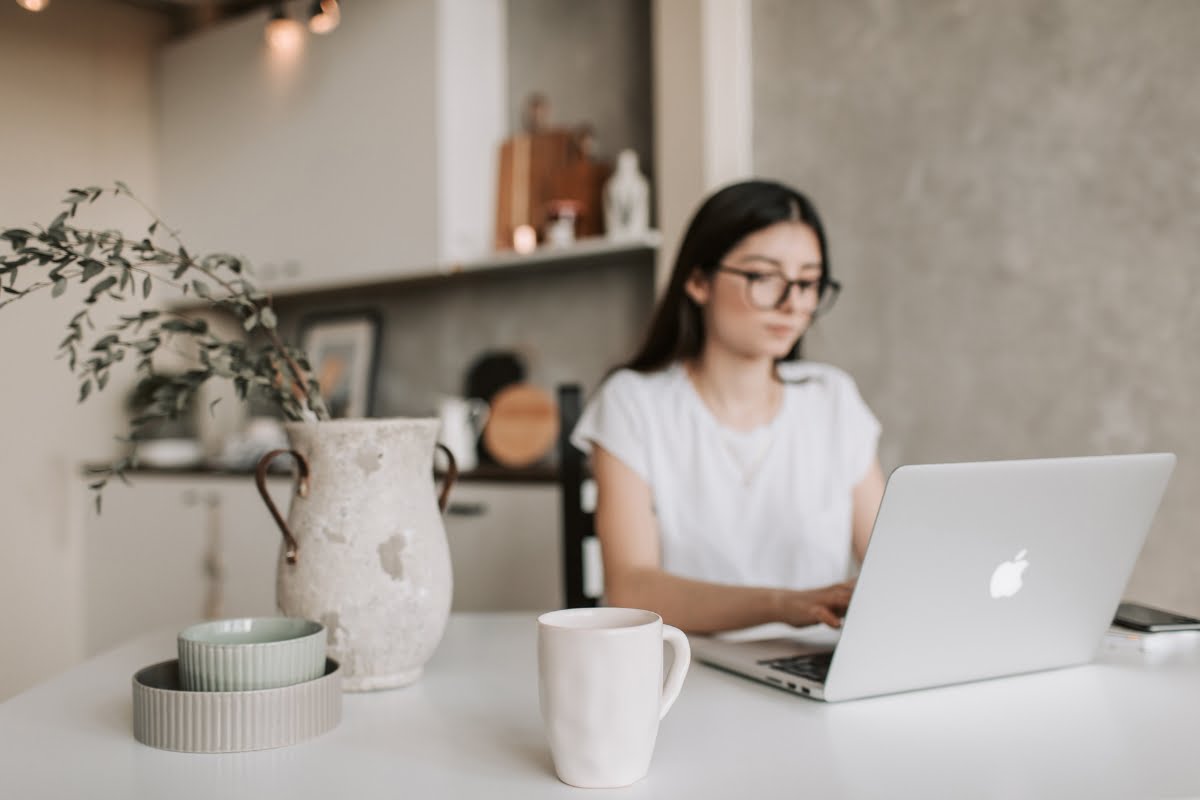 A woman with long hair and glasses is working on a laptop at a table, with a white mug and a vase with branches in the foreground. The cozy setup feels like one of the best places to work remotely in Atlanta, as the background reveals a kitchen adorned with various items on the counter.