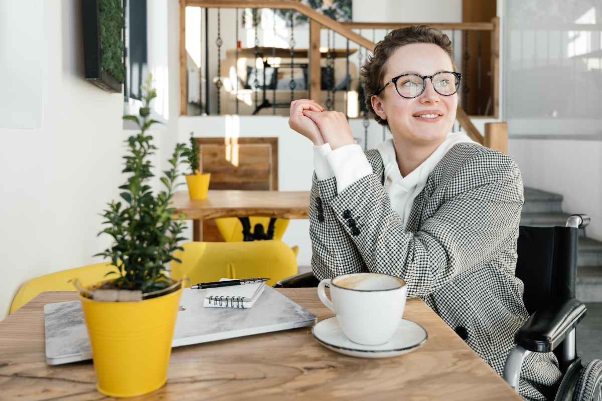 Woman in a checkered blazer sits at a café table with a notebook and coffee cup, smiling and looking to the side. A small plant in a yellow pot is in the foreground, capturing the essence of one of the best places to work remotely in Atlanta.