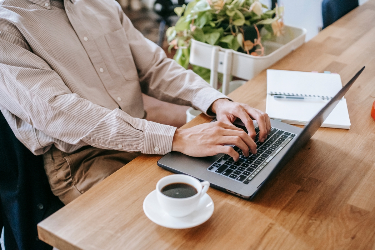 A person types on a laptop at a wooden desk with a cup of coffee, an open notebook, and a plant nearby, perhaps researching the best places to work remotely in Atlanta.