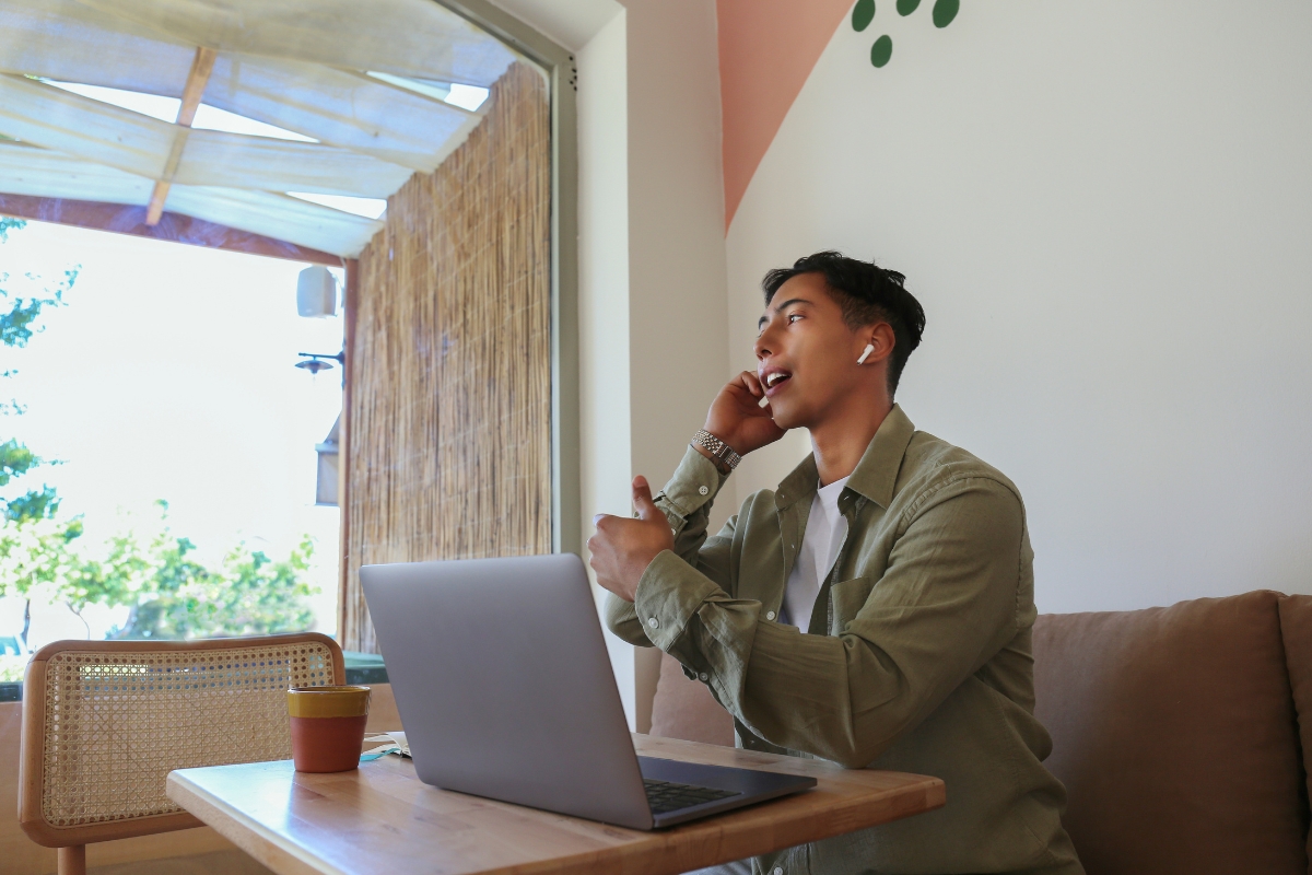 A person wearing a green shirt and earbuds sits at a wooden table using a laptop and talking on a smartphone in one of the best places to work remotely in Atlanta, bathed in natural light from the nearby window.