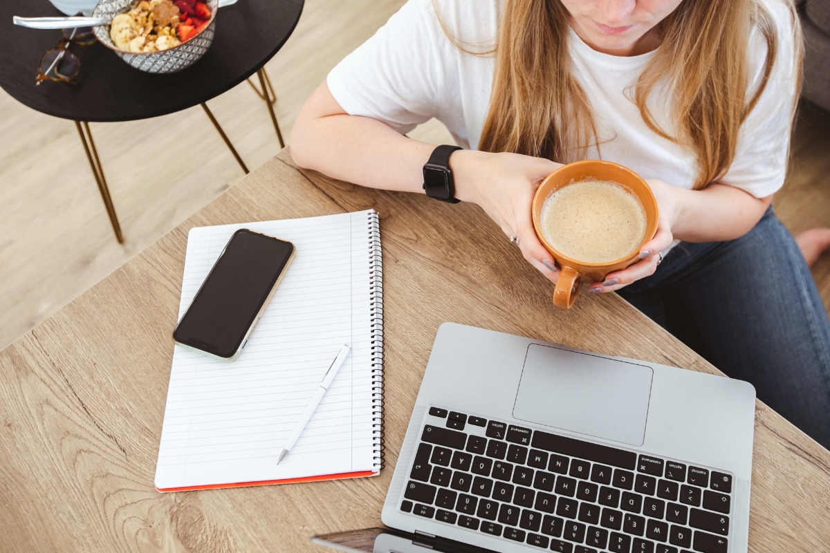 A person sits at a desk with a laptop, holding a cup of coffee. Nearby, a notebook with a pen and smartphone rest on the desk, alongside a bowl of food on the table. This setup resembles one you'd find in the best places to work remotely in Atlanta.