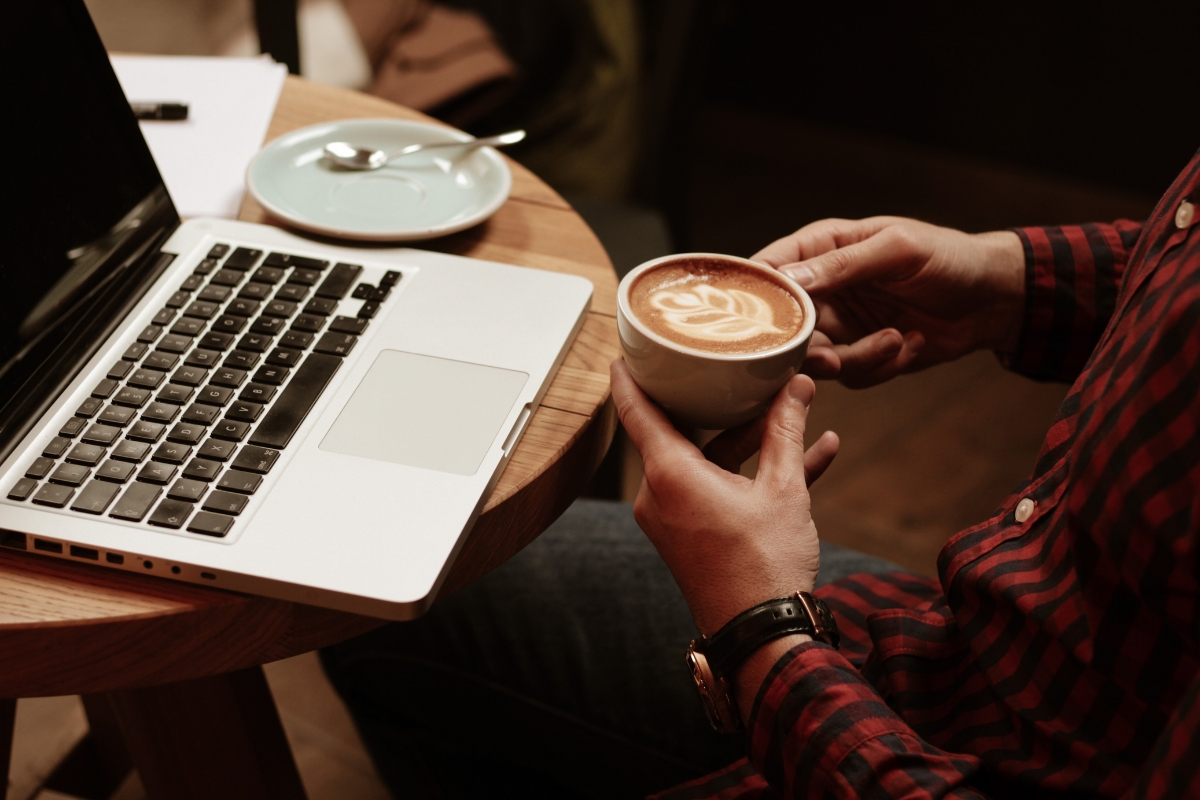 A person in a red plaid shirt holds a latte with latte art while sitting at a table with an open laptop, a small plate, and a spoon, perhaps exploring the best places to work remotely in Atlanta.