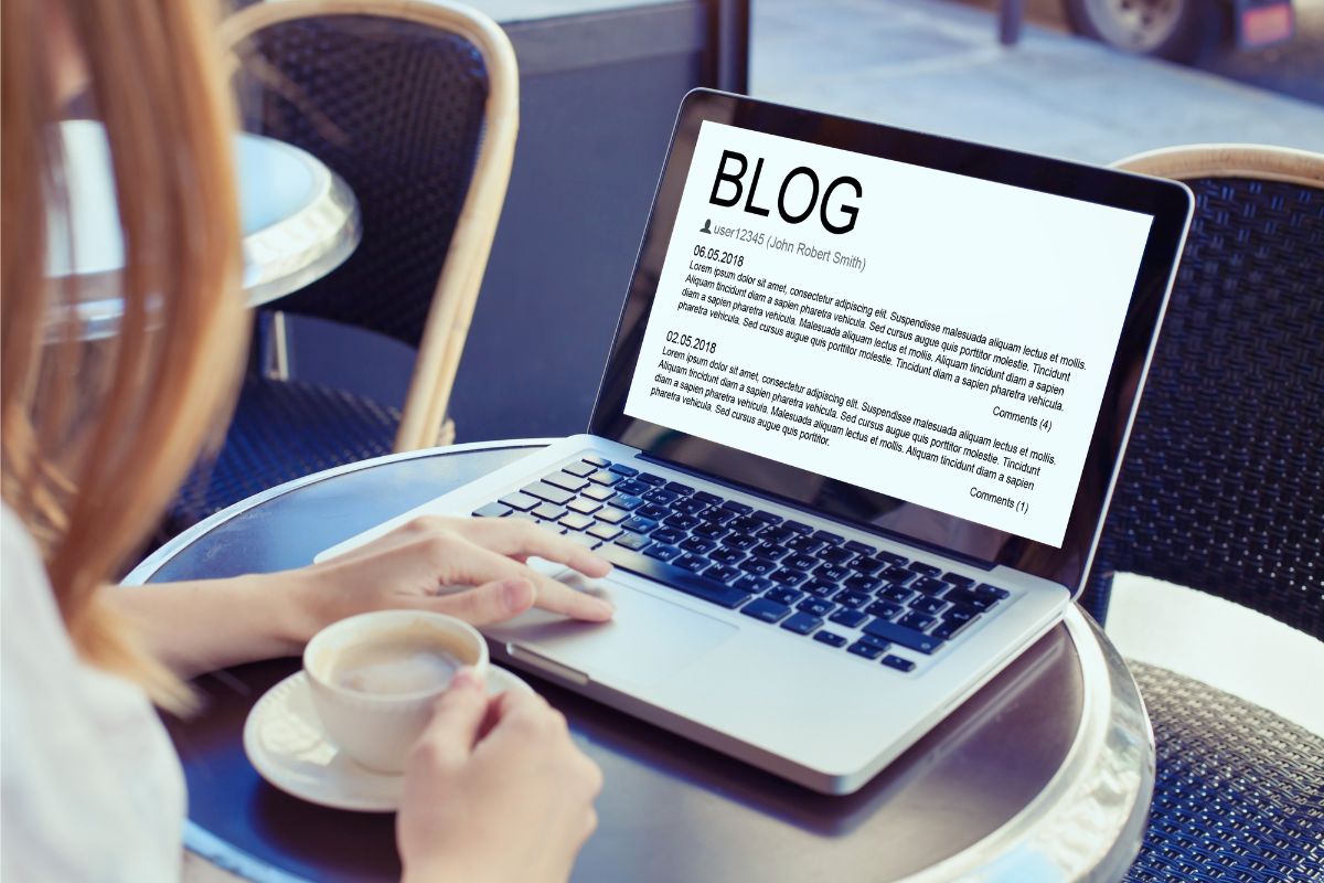 A person reading a blog post titled "How To Write A Blog Post" on their laptop screen while holding a cup of coffee at a cozy café table.