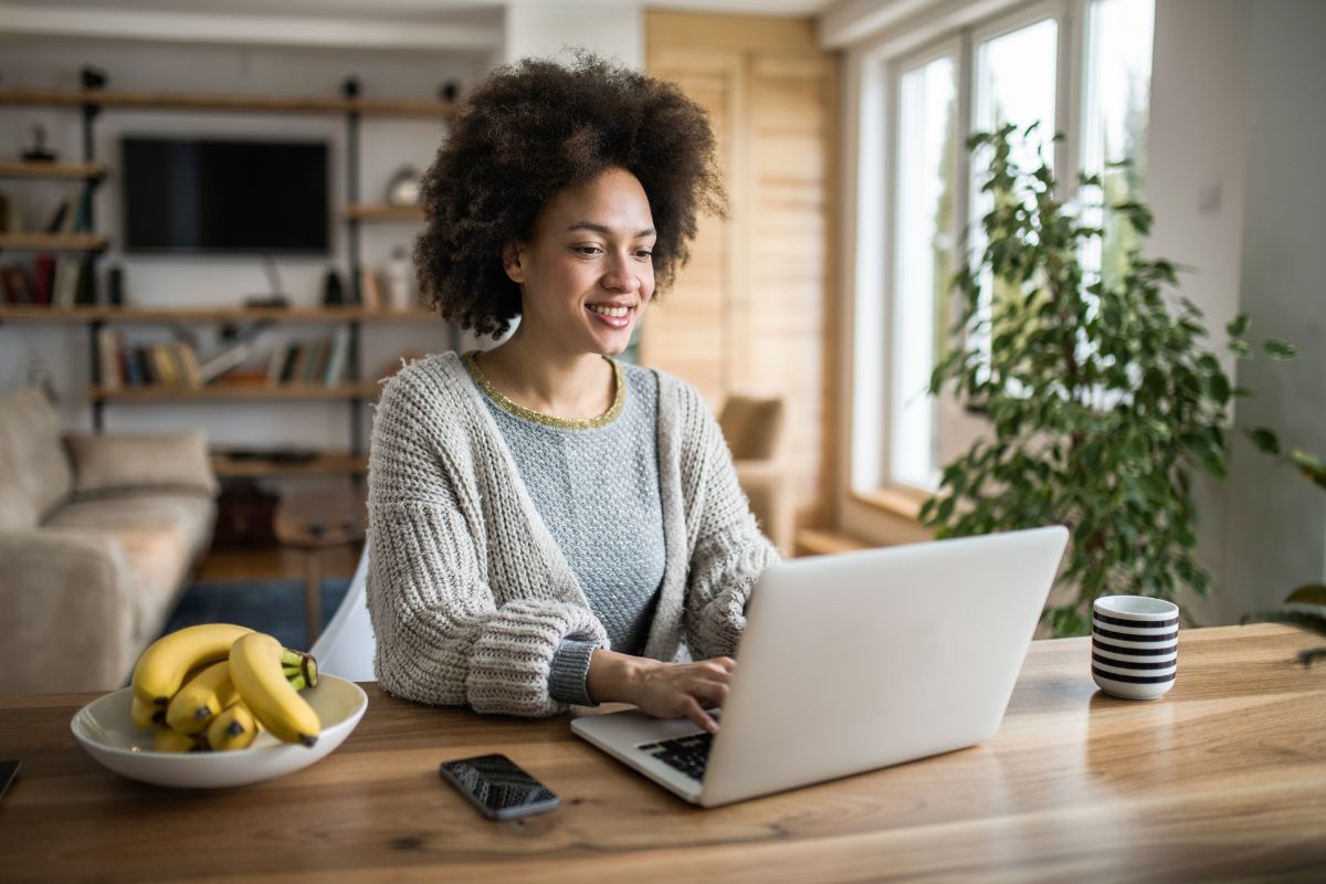 A woman with curly hair works on a laptop at a wooden table in a bright living room, perhaps drafting her next piece on how to write a blog post. There is a bowl of bananas, a smartphone, and a striped mug on the table. Shelves and a plant are in the background.