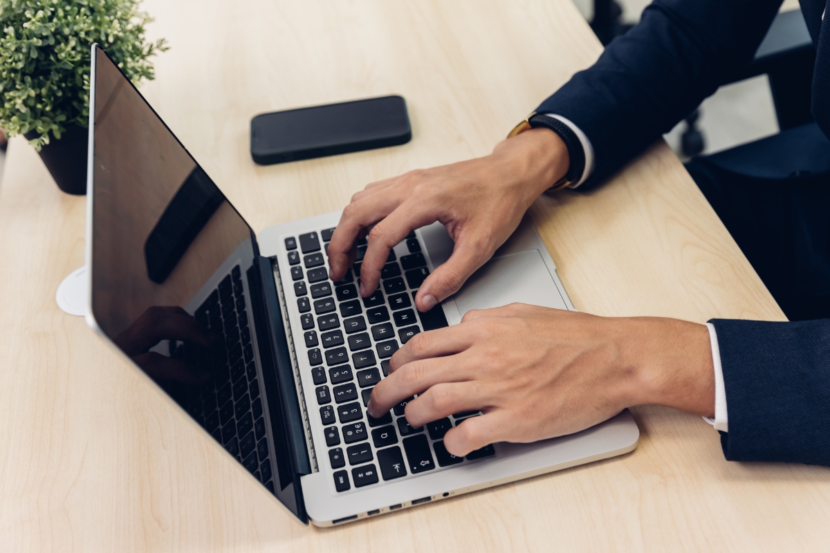 A person in a suit types on a laptop placed on a wooden desk with a smartphone nearby, pondering the question, "can Google detect AI content?" A small potted plant adds a touch of greenery to the serene workspace.