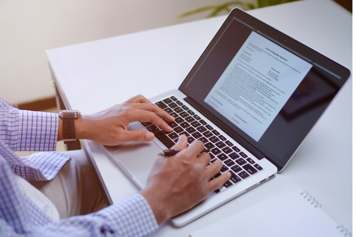 Person in a blue-checkered shirt typing on a laptop displaying a document about "can Google detect AI content," working at a white desk with a notebook nearby.