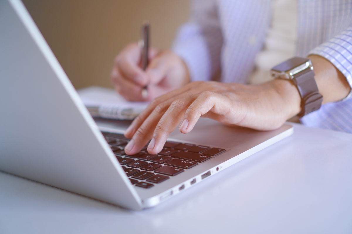 A person wearing a striped shirt uses a laptop while holding a pen. Only their hands and part of the torso are visible, perhaps contemplating, "Can Google detect AI content?