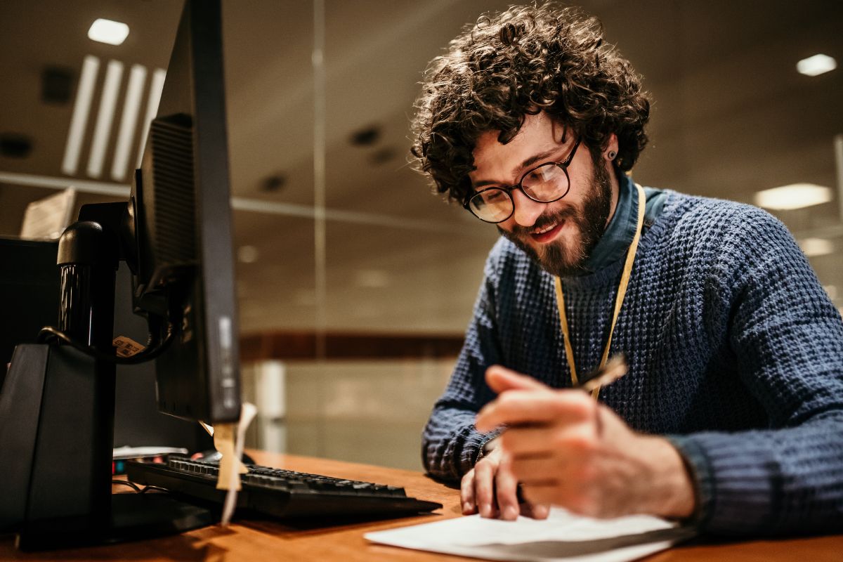 A person with curly hair and glasses, wearing a blue sweater, works at a desk with a computer and documents, holding a pen. They appear to be engaged in creating or organizing ChatGPT prompts.