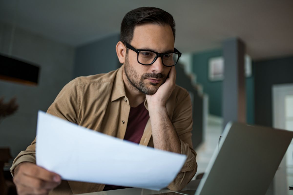 A man wearing glasses and a beard looks at a laptop screen while holding a piece of paper in his hand. He appears focused, likely drafting ChatGPT prompts, seated at a desk in a home setting.