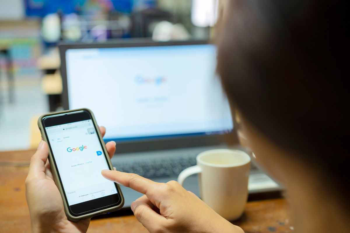 A person uses a smartphone to search on Google for "How To Do Keyword Research," with a laptop displaying the Google homepage in the background and a cup beside it.