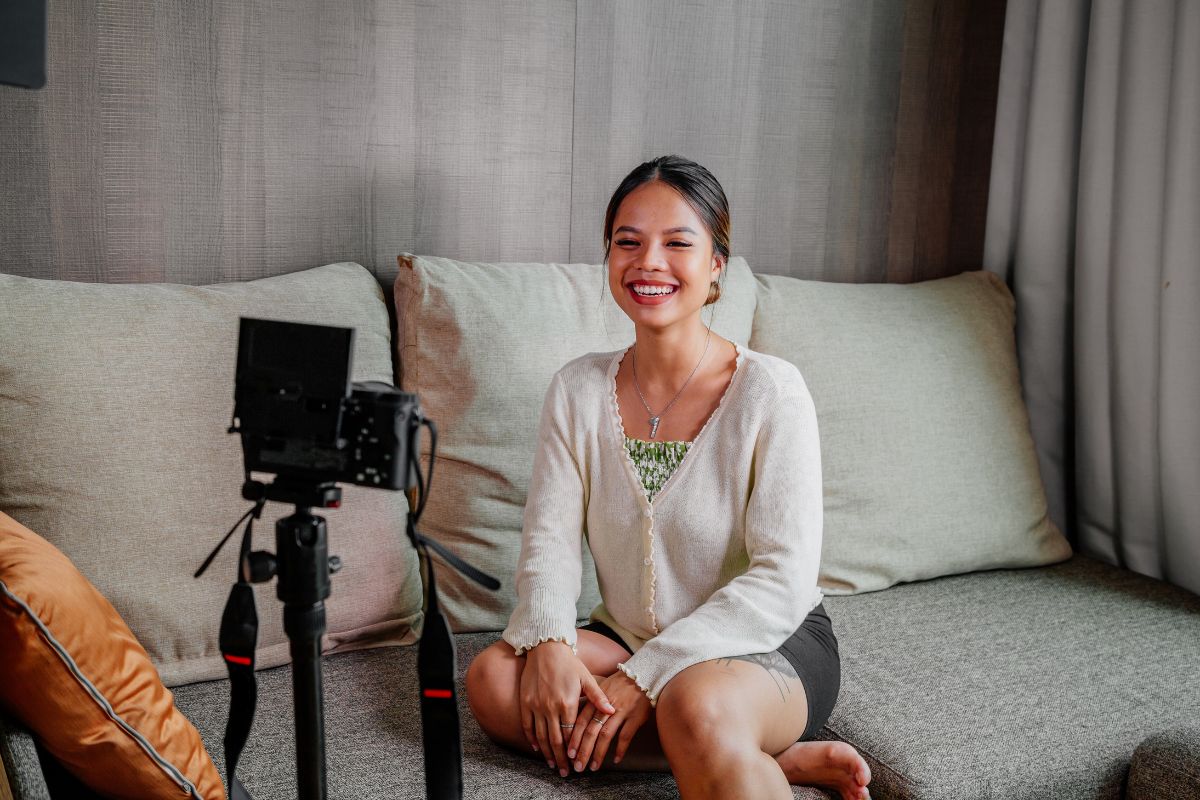 A smiling person sits on a couch in front of a camera setup, possibly recording video testimonials.