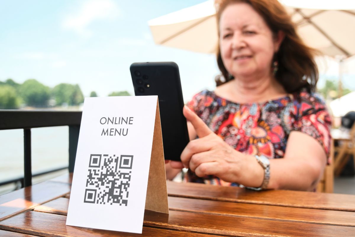 A woman scans a QR code labeled "Online Menu" with her phone at an outdoor dining area, seamlessly integrating modern technology into her dining experience. Curious diners often wonder, what are QR codes? These nifty tools provide easy access to digital menus and more with just a scan.