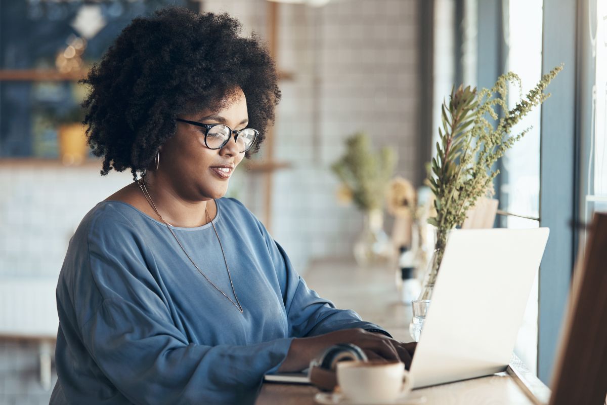 A person with curly hair and glasses works on a laptop at a cafe, crafting the perfect nurture campaign, with a cup of coffee and plants on the wooden counter.